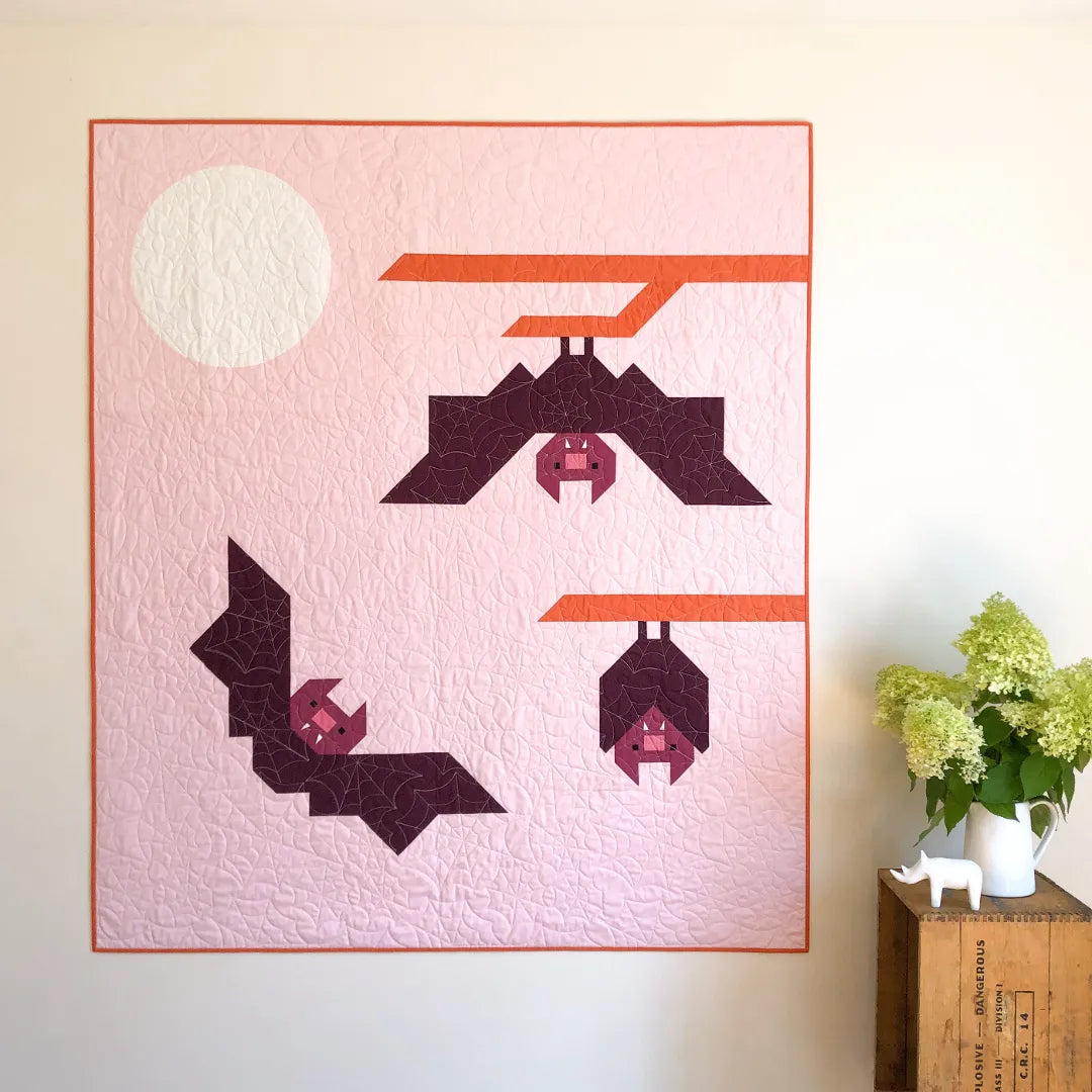 The Bat Buddies Quilt Pattern by Apples & Beavers features modern geometric bats hanging from orange branches on a pale pink background with a white full moon—perfect for adding spooky charm to your Halloween décor.