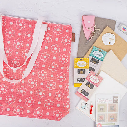 A Big Daisy Project Bag in pink with white daisies is displayed on a light surface, surrounded by quilting accessories like needles, keychains, fabric pieces, a seam ripper, quilt blocks, and stitch cards.