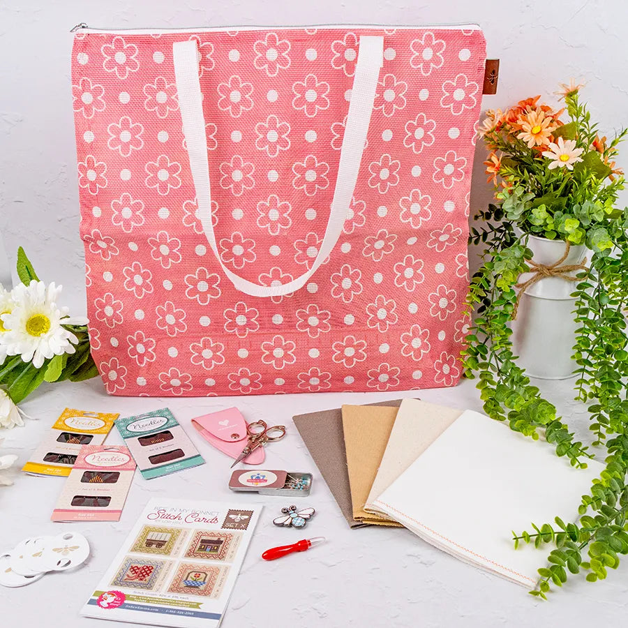 A Big Daisy Project Bag in pink with white daisy patterns is surrounded by quilting and cross stitch supplies, scissors, fabric, embroidery threads, tools, and faux flowers on a white background.