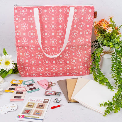 A Big Daisy Project Bag in pink with white daisy patterns is surrounded by quilting and cross stitch supplies, scissors, fabric, embroidery threads, tools, and faux flowers on a white background.