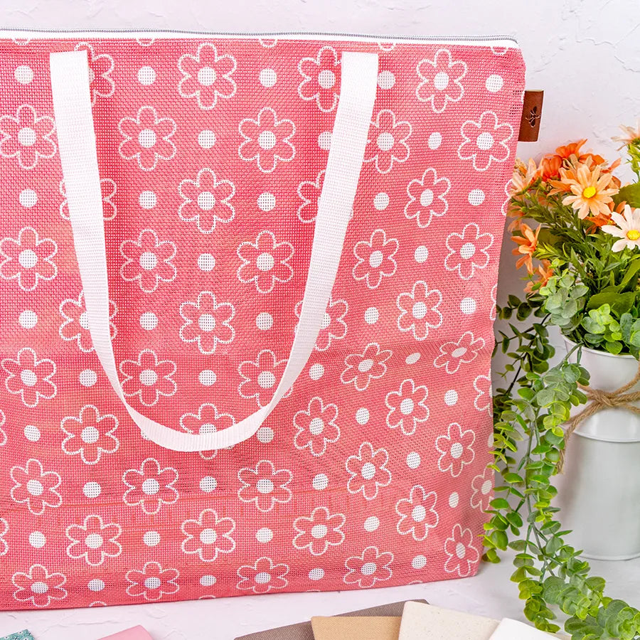 The Big Daisy Project Bag, a pink quilted bag with white handles and a white daisy pattern, sits beside yellow and orange flowers in a white vase—ideal for storing your quilting accessories.