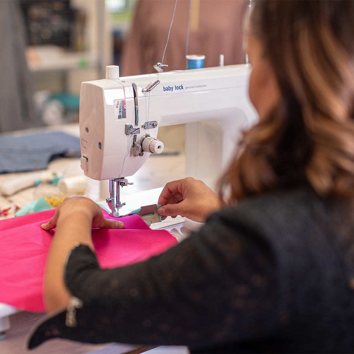 A beginner sewing enthusiast uses the Baby Lock Genuine Collection Pixie Playdate machine to stitch bright pink fabric, carefully guiding the material by hand in a workshop setting.