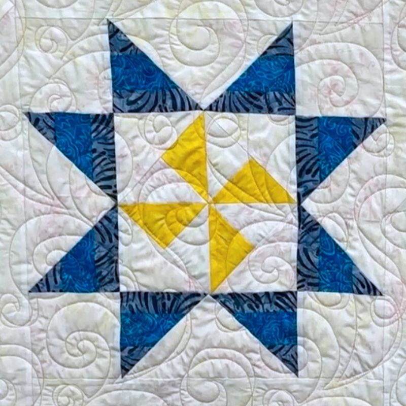 A quilt block featuring a star pattern with blue triangles, white triangles, and a central yellow pinwheel design, all set on a white background with quilted swirling lines.