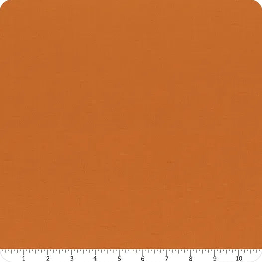 A square of Riley Blake Confetti Cotton™ Amber Solid Fabric with a subtle texture is shown, featuring a white ruler along the bottom from 0 to 12 inches. The fabric’s rounded corners make it perfect for quilting and other cotton projects.