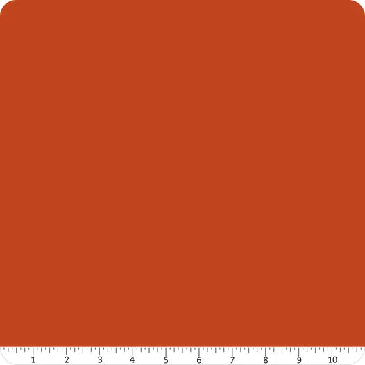 A square swatch of Confetti Cotton™ Rusty Fabric Yardage by Riley Blake, in solid reddish-brown, features rounded corners and a white ruler along the bottom edge with 0–10 inch markings. Ideal for cotton quilting projects.