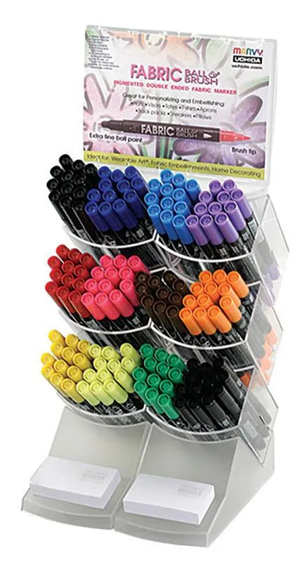A clear acrylic display holds rows of colorful United Notions Marvy Fabric Ball & Brush Markers organized by color, with a "Fabric Brush" sign at the top and two white pads at the bottom.