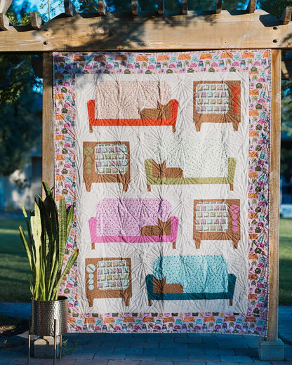 The Couch Potatoes Quilt Pattern by Checker Distributor, featuring vibrant sofas and armchairs, hangs outdoors on a wooden pergola with a potted snake plant in front.