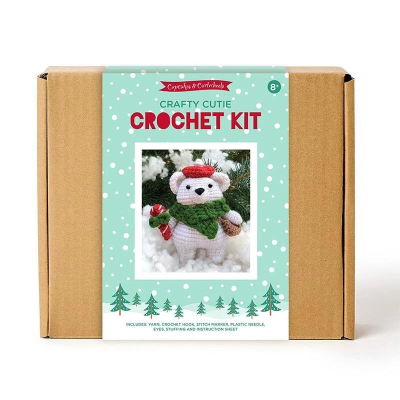 The United Notions Crafty Cutie Polar Bear Crochet Kit comes in a box with an image of a bear in a green scarf and red hat, holding a candy cane amid snow and trees—ideal for beginners seeking a festive crochet project.