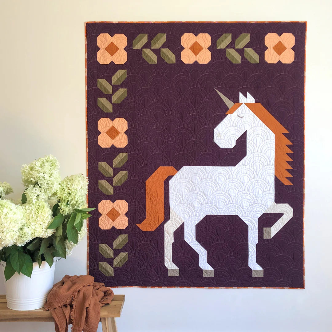 The Unicorn Garden Quilt Pattern by Apples & Beavers features a whimsical white unicorn with an orange mane on a purple background, bordered by orange and green patchwork flowers and leaves, with a white vase of flowers on a wooden bench in front.