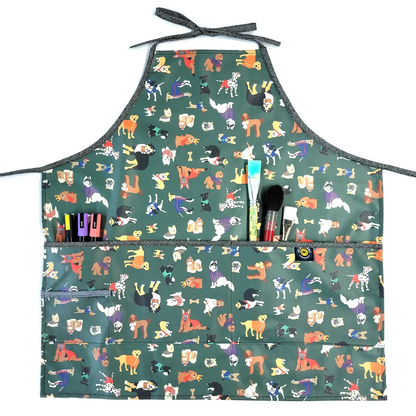 The Full Apron in Laminated Cotton, Yard Dogs in Green, features colorful dog illustrations on a green background and several front pockets—ideal for holding pens, markers, or brushes during creative gardening or indoor tasks.