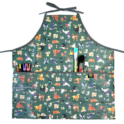 The Full Apron in Laminated Cotton, Yard Dogs in Green, features colorful dog illustrations on a green background and several front pockets—ideal for holding pens, markers, or brushes during creative gardening or indoor tasks.