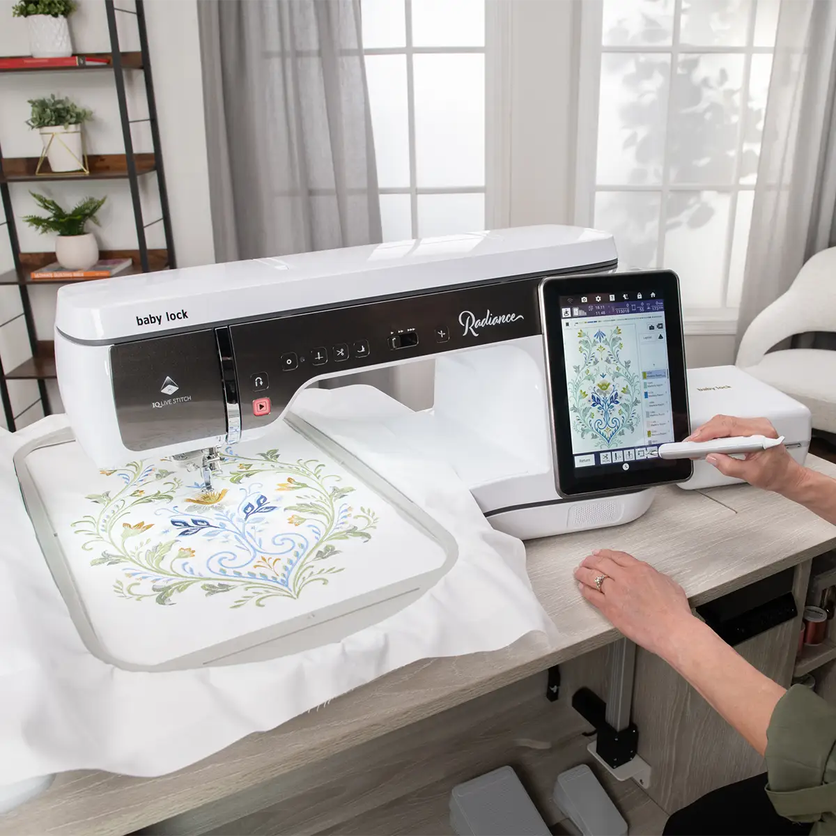 A person uses a stylus to operate the IQ Designer touchscreen on the Baby Lock Embroidery 3 Pixie Playdate, which stitches a colorful floral pattern onto white fabric in a bright, modern room.