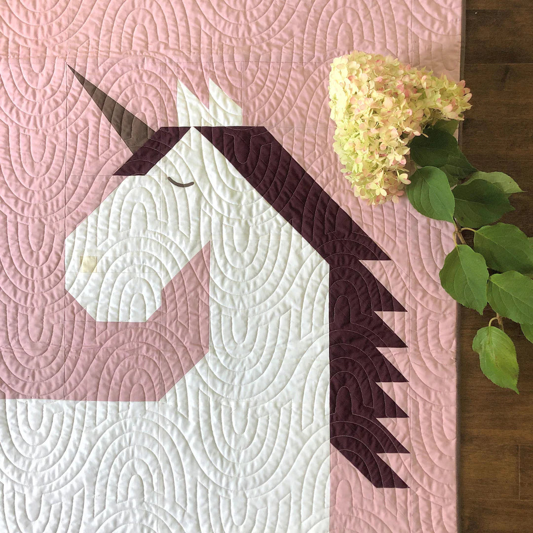 The Unicorn Garden Quilt Pattern by Apples & Beavers features a geometric white unicorn with a dark mane and horn on a pink background, accented by pale pink hydrangeas and green leaves in the top right corner.