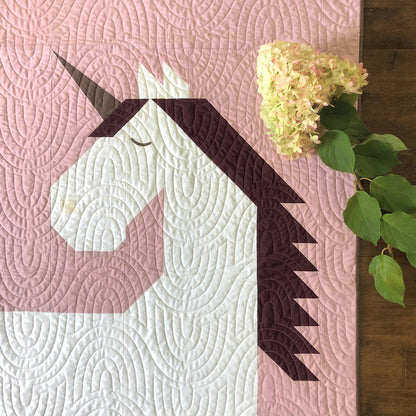 The Unicorn Garden Quilt Pattern by Apples & Beavers features a geometric white unicorn with a dark mane and horn on a pink background, accented by pale pink hydrangeas and green leaves in the top right corner.