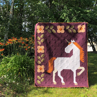 The Unicorn Garden Quilt Pattern by Apples & Beavers features a white unicorn with an orange mane and horn on maroon, accented with flowers and leaves—a whimsical design perfect for garden-inspired quilts.
