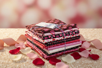 The Andover Heart and Soul Half Yard Bundle, featuring pink, red, and black heart-patterned fabrics tied with string, is arranged on a lace-covered surface surrounded by rose petals and ribbon against a soft pink background.