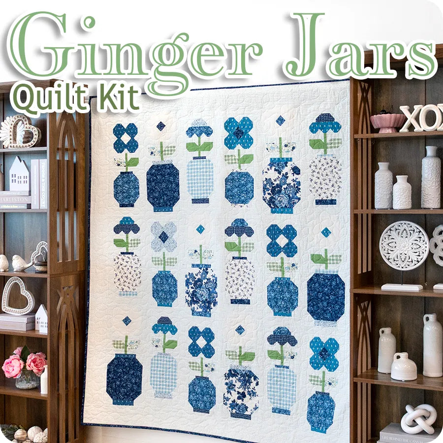 A blue and white quilt with flower designs in patterned jars hangs between two shelves with decor. “Blueprint Quilt Kit” by United Notions is featured, made with United Notions fabrics and inspired by the Blueprint Quilt Pattern.