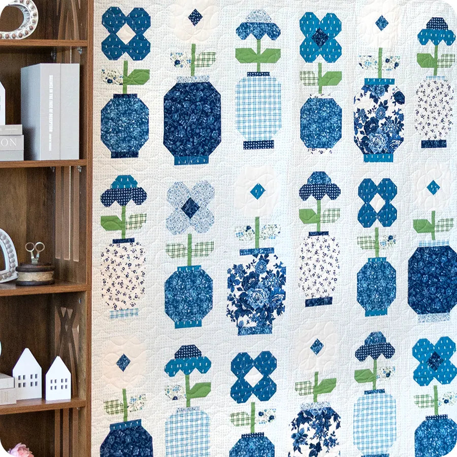A Blueprint Quilt Kit by United Notions, made with blue and white fabrics in flower and vase motifs, hangs on the wall. A wooden shelf with books and house ornaments sits to the left for added charm.