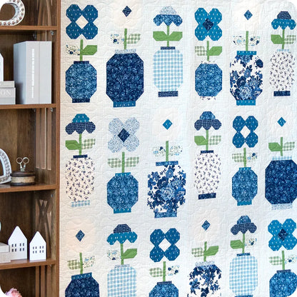 A Blueprint Quilt Kit by United Notions, made with blue and white fabrics in flower and vase motifs, hangs on the wall. A wooden shelf with books and house ornaments sits to the left for added charm.