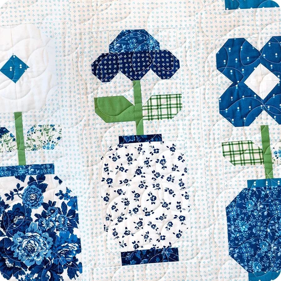 The Blueprint Quilt Kit by United Notions features quilted fabric inspired by Camille Roskelley, with three blue and white floral patterns in vases, unique prints, green stems and leaves, and a light background accented with blue dots.