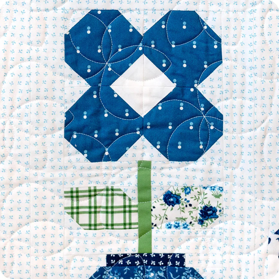 The Blueprint Quilt Kit by United Notions includes a quilted block inspired by the Ginger Jars Quilt Pattern, showcasing a blue flower with a white diamond center, green and printed leaves, and a blue vase on a light patterned background.