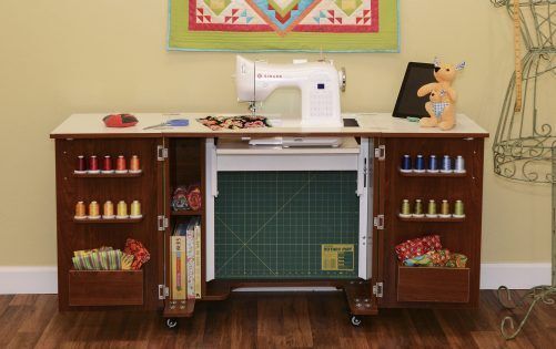 A sewing machine sits on an Arrow Companies, LLC Kangaroo Bandicoot II Sewing Cabinet, with shelves holding colorful thread, fabric, and supplies. An open compartment shows a cutting mat. A stuffed deer toy and quilt appear in the background.