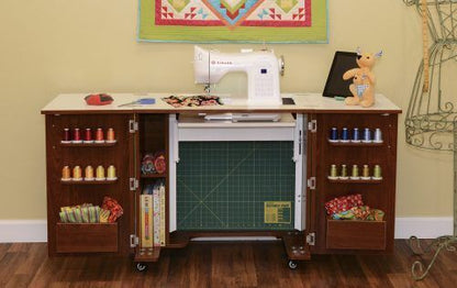 A sewing machine sits on an Arrow Companies, LLC Kangaroo Bandicoot II Sewing Cabinet, with shelves holding colorful thread, fabric, and supplies. An open compartment shows a cutting mat. A stuffed deer toy and quilt appear in the background.