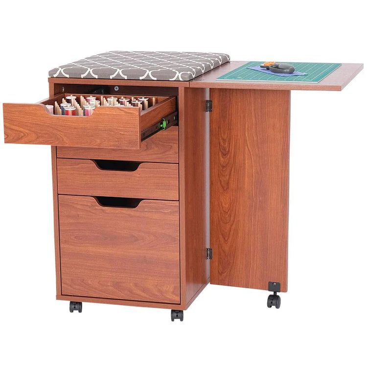 The Kiwi Multi-Use Cabinet offers a cushioned top, open side table, large cutting surface, open drawer with thread spools, two closed drawers, and sewing tools on the extended tabletop—all on wheels for easy mobility.