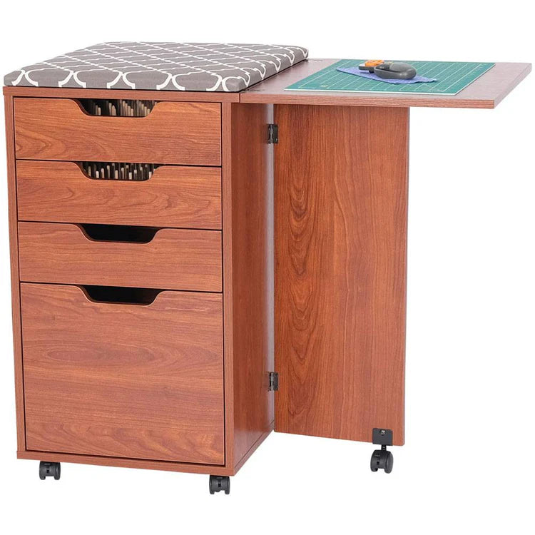 The Kiwi Multi-Use Cabinet features three drawers, a cushioned top, a partially open side extension, wheels for mobility, and a green cutting mat with sewing tools on the extended cutting surface.