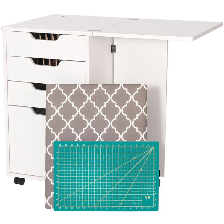 The Kiwi Multi-Use Cabinet is versatile sewing furniture in white, with open and closed drawers, a fold-out tabletop, a gray and white cushion, and a green grid-lined cutting surface in front.