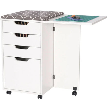 The Kiwi Multi-Use Cabinet features a gray cushioned top, three drawers, a large compartment, and a fold-out cutting surface. A green cutting mat and rotary cutter are placed on the extended section of this white rolling craft table.
