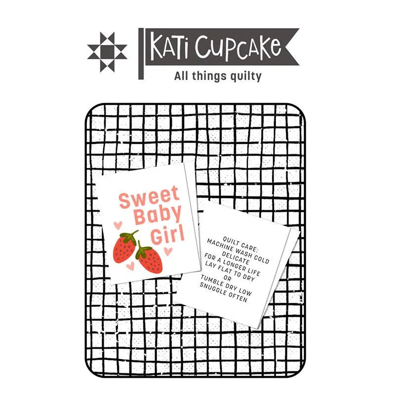 A graphic with the Kati Cupcake logo above two cards on a black-and-white grid background features the Sweet Baby Girl Sew in Labels 3 pack, showing a strawberry "Sweet Baby Girl" card and quilt care instructions for handmade creations.