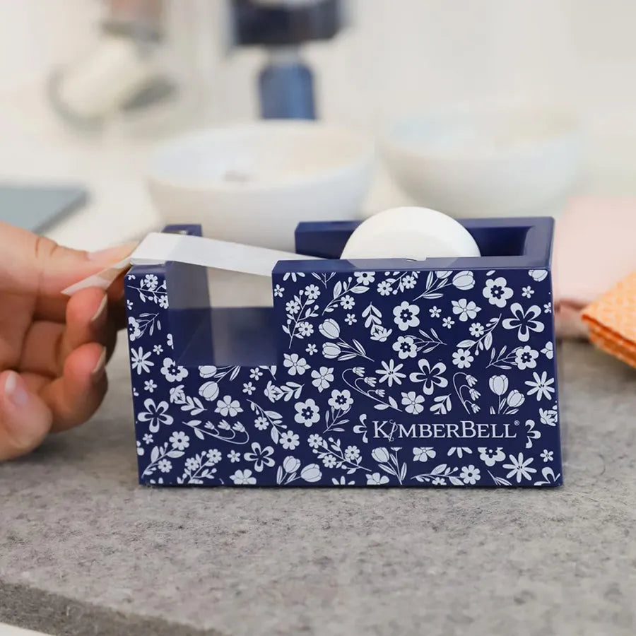 A hand pulls Kimberbell Paper Tape from the Kimberbell Navy Floral Paper Tape Dispenser by Checker Distributor, featuring white floral patterns and the brand name, perfect for embroidery projects, on a gray surface.