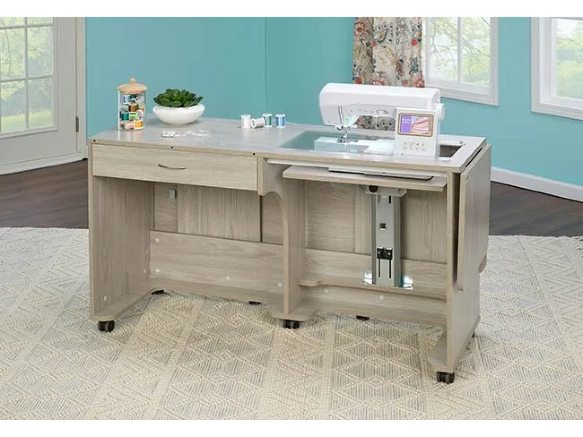 The Koala Quilters Vision Sewing and Companion Chest in White and Gray, featuring a sewing machine on top, small drawer, storage compartments, and rolling casters, stands in a bright room with teal walls and patterned carpet.