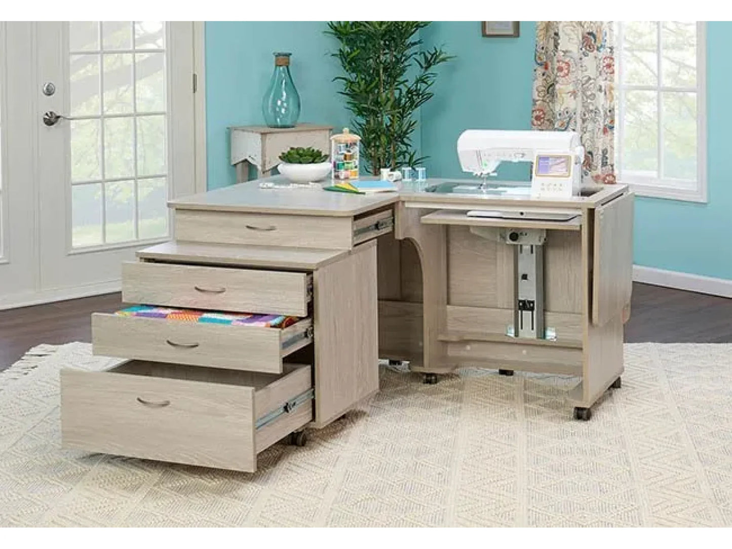 The Koala Quilters Vision Sewing and Companion Chest in White and Gray is a stylish sewing machine cabinet with open drawers displaying colorful fabric, placed by a pull-out platform in a bright, blue-walled room with large windows and a plant.