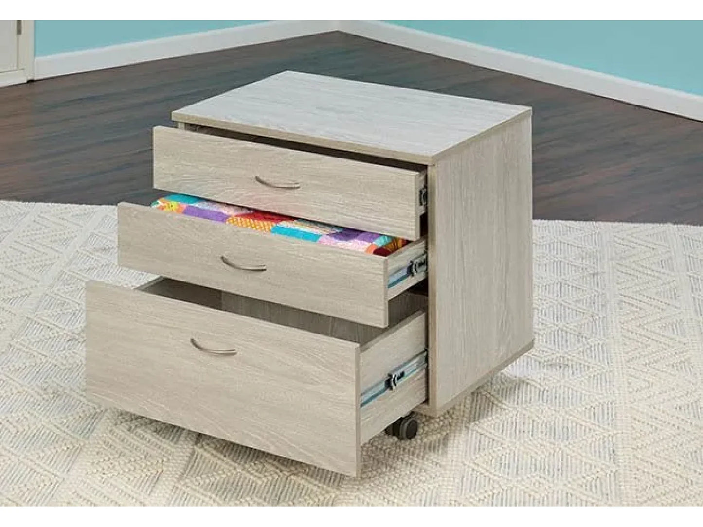 The Koala Quilters Vision Sewing and Companion Chest in White and Gray with three drawers on wheels sits on a textured rug. Two drawers are open, showing a colorful quilt. The cabinet is in a blue-walled room with dark floors.
