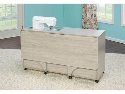 The Koala Quilters Vision Sewing and Companion Chest in White and Gray, with a drop-leaf extension and caster wheels, is set in a bright room with blue walls, white trim, large windows, and patterned curtains.