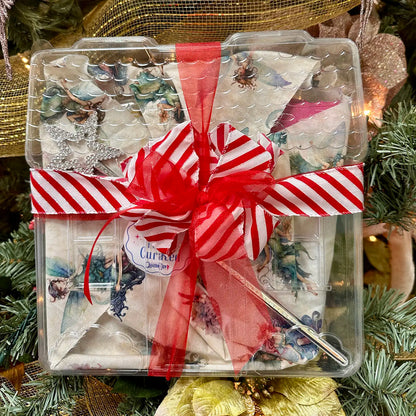 The Quilted Joy Limited Edition Quilt Fairy Magic Mystery Box features angel-themed napkins, a silver star, and a fork in a festive gift box with a red and white bow—an enchanting holiday surprise under the Christmas tree.