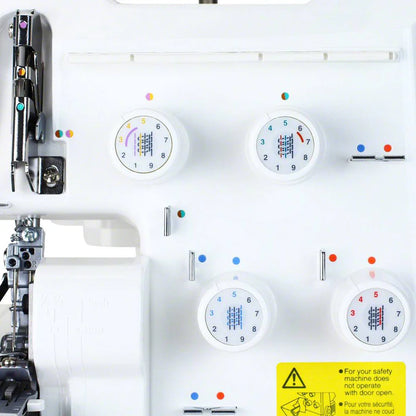Close-up of a Juki America Pearl Line MO-655 2/3/4/5 Thread Serger control panel, featuring four colored dials, metal components, and a yellow safety warning label at the bottom right.