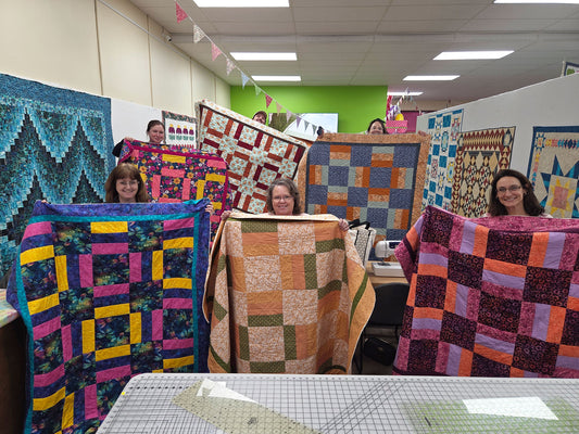 Five women smile and display colorful quilts in a craft room during My First Quilt Class Night by Quilted Joy, surrounded by bunting, quilts, and tables filled with quilting tools—capturing a lively beginner quilting experience.