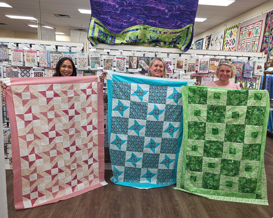 Four women stand indoors, each holding a colorful quilt with geometric patterns in pink, blue, and green. Behind them hang samples from the Quilted Joy "My Next Quilt Class: Half Square Triangles - Night" class and half square triangles.
