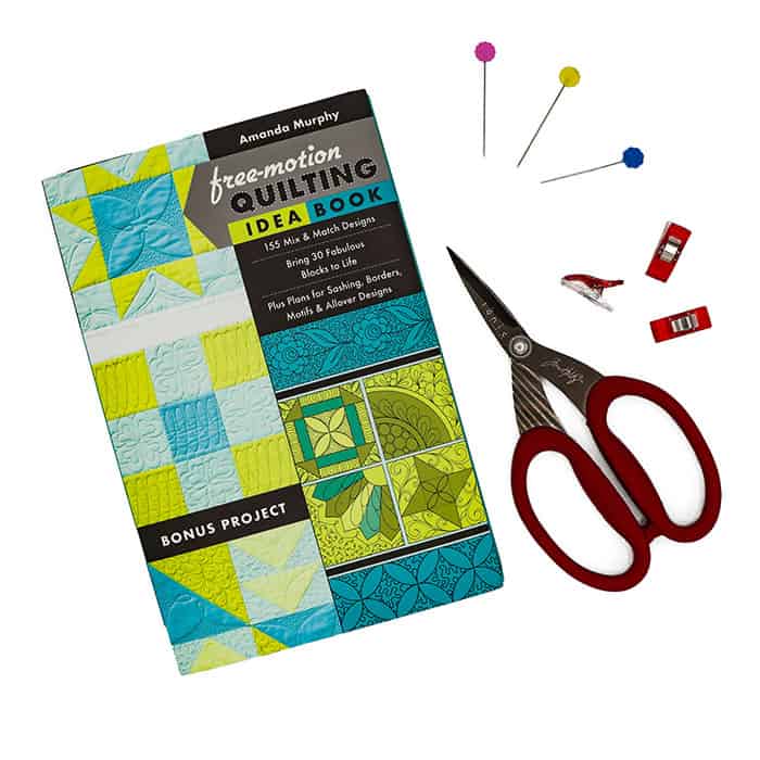 A quilting idea book, a pair of red-handled scissors, three sewing pins with colorful heads, and two red sewing clips are arranged on a white background.