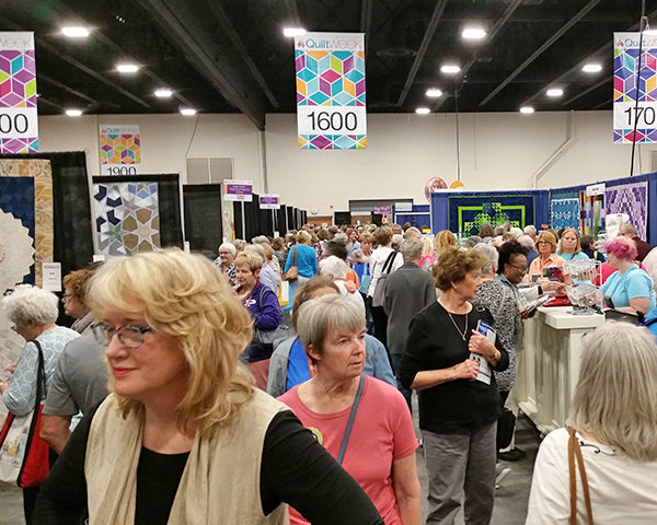 A large group of mostly older adults walk and chat at the indoor "Bus To Paducah 2026" quilt show by Quilted Joy, surrounded by colorful quilts, booths with quilting goodies, and bright numbered signs hanging overhead.