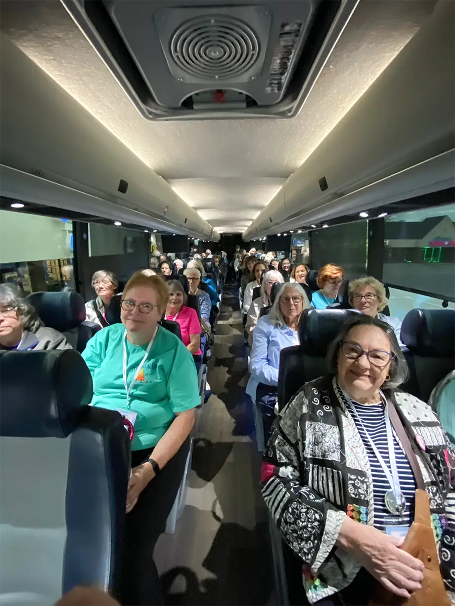 A group of older adults ride Quilted Joy’s Bus To Paducah 2026, relaxing on a well-lit coach with spacious seats and large windows, excited for AQS show admission and shopping at Hancock’s of Paducah.