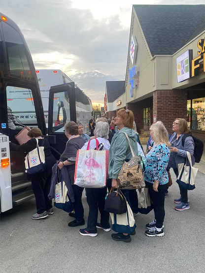 A group of women with Quilted Joy tote bags filled with quilting supplies wait on the sidewalk to board the Bus To Paducah 2026, parked outside shops under a cloudy sky.