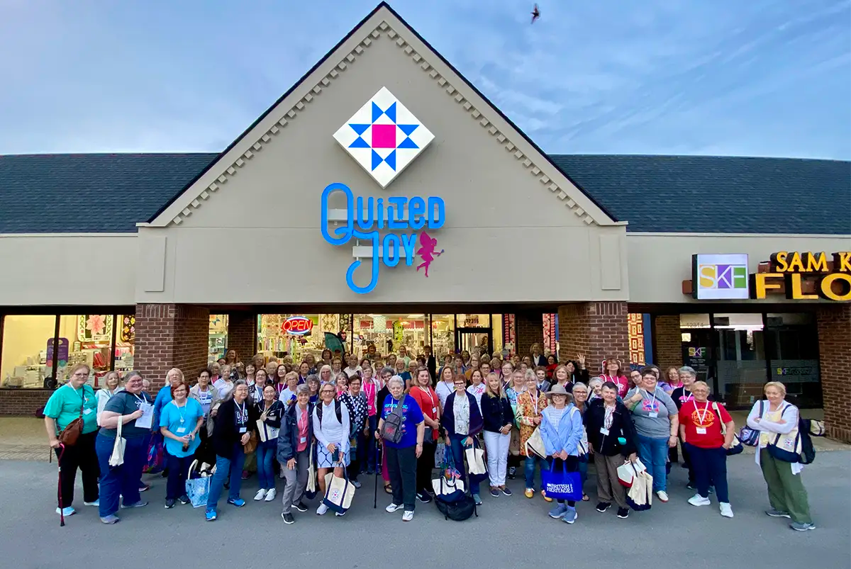 A large group smiles in front of Quilted Joy, holding Bus To Paducah 2026 tote bags filled with quilting goodies, outside the shop in a lively plaza under a clear blue sky.