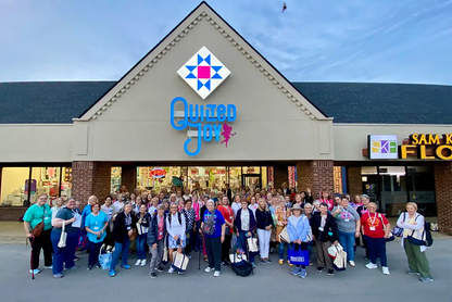 A large group smiles in front of Quilted Joy, holding Bus To Paducah 2026 tote bags filled with quilting goodies, outside the shop in a lively plaza under a clear blue sky.