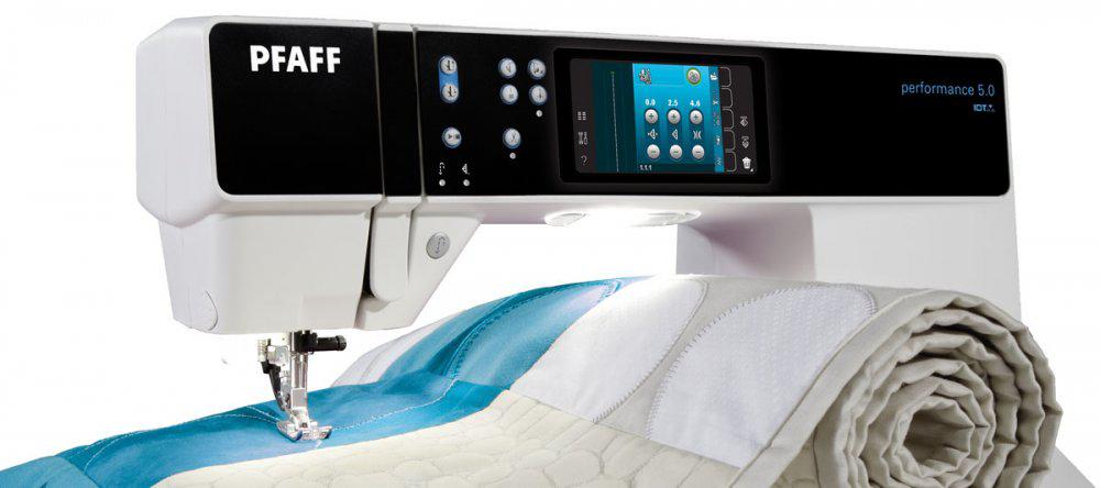 A close-up of the PRE-LOVED Pfaff Performance 5.0 sewing machine by Quilted Joy in use, stitching a white and blue quilt, with its digital screen displaying various controls.