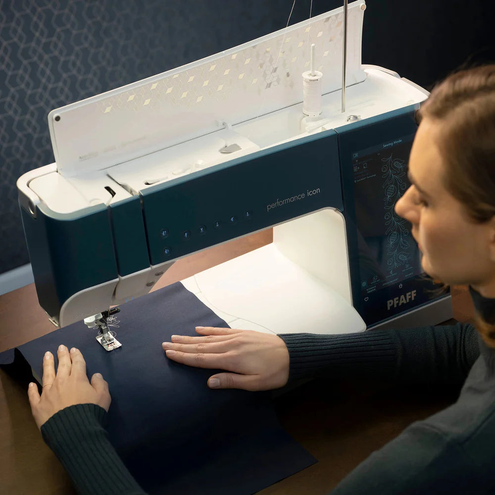 A person guides dark fabric through a blue and white Pfaff Pixie Playdate sewing machine on a wooden table, focusing on stitching.