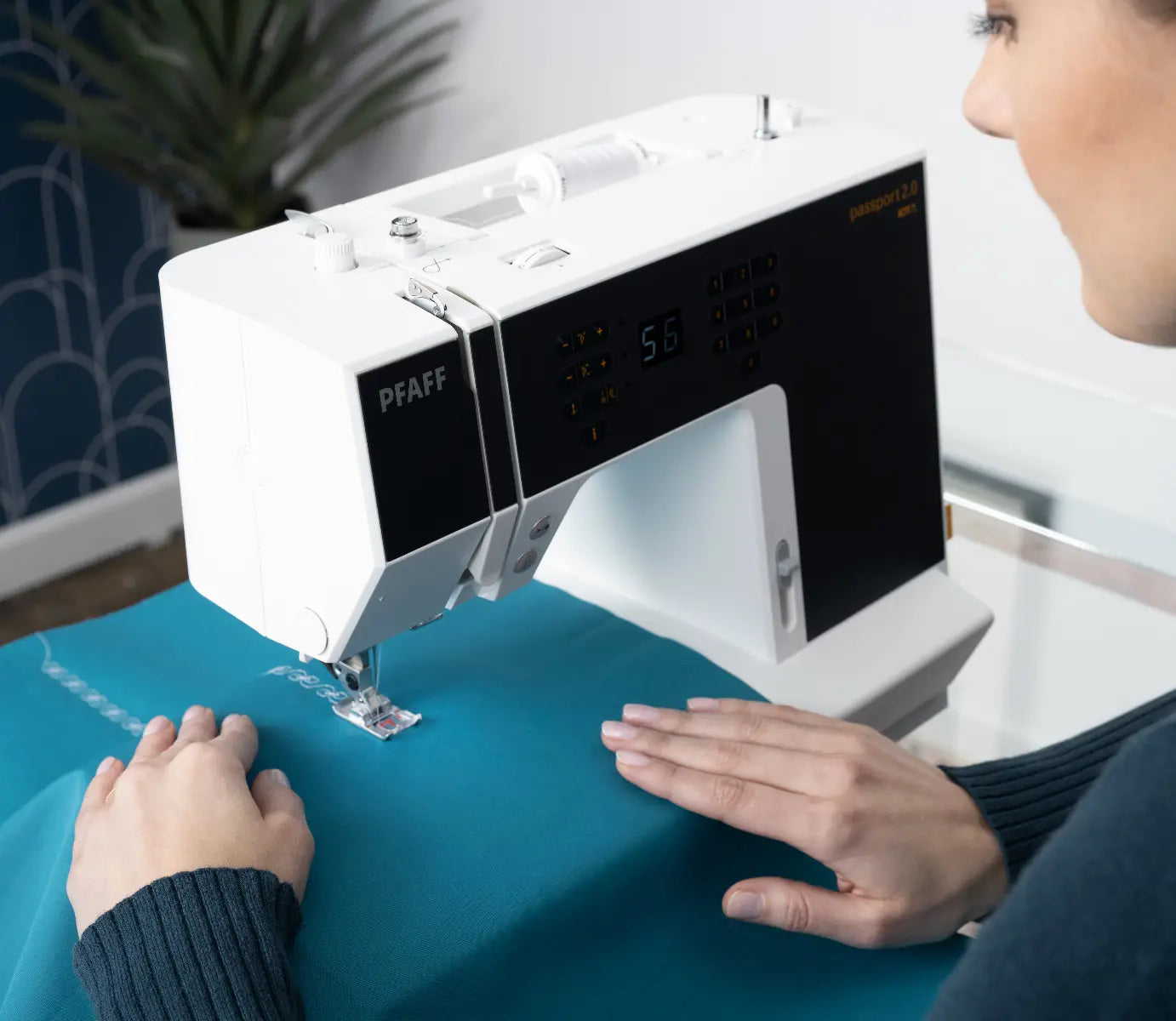 A person uses a white and black PFAFF sewing machine to stitch blue fabric, guiding it carefully with both hands on a glass table.